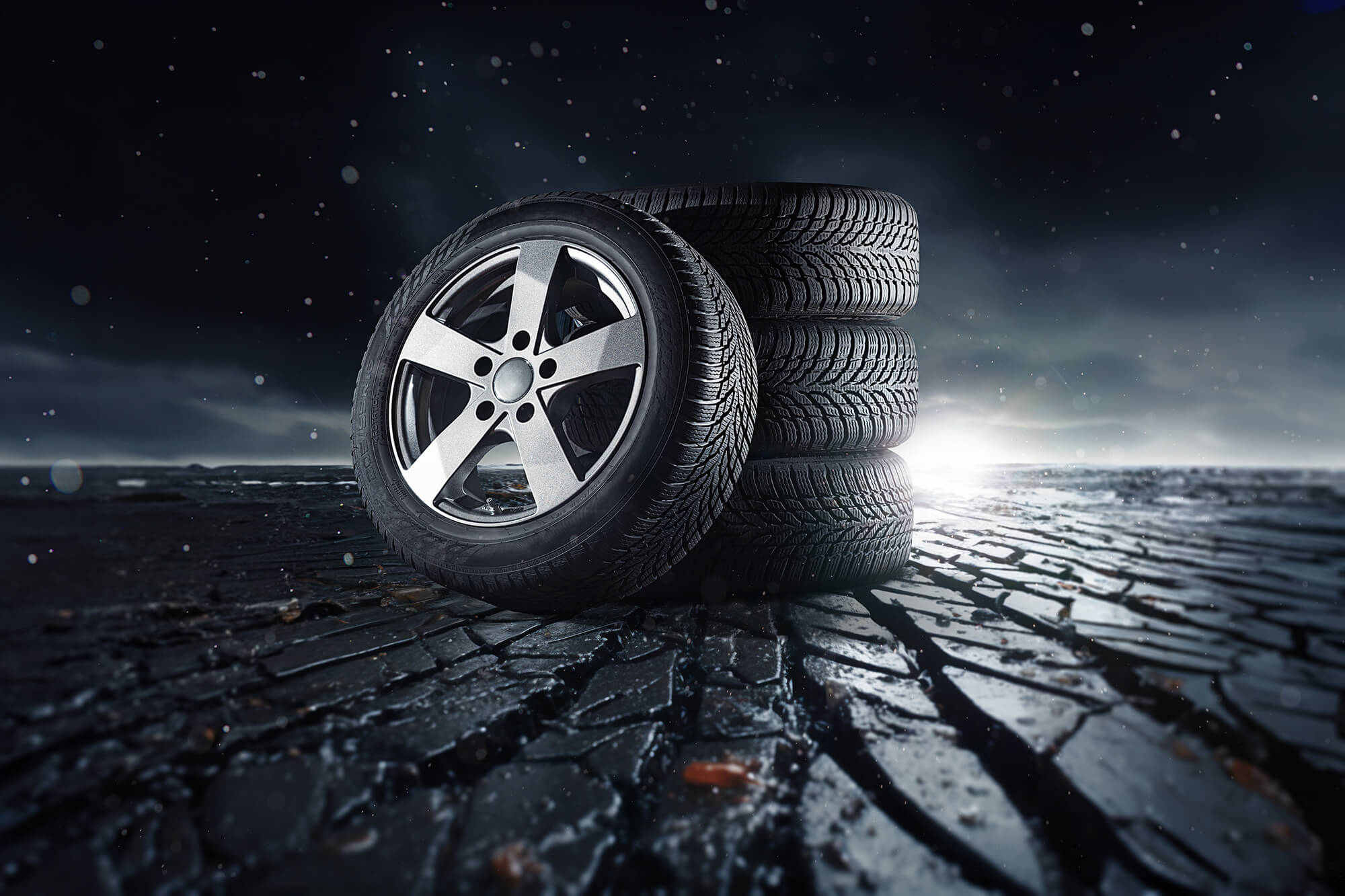 Four car tires on a cracked surface under a dark, dramatic sky, one standing upright and three stacked behind it.